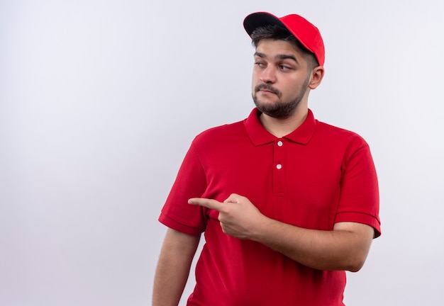 Repartidor joven con uniforme rojo y gorra mirando a un lado con expresión escéptica apuntando con el dedo índice hacia un lado