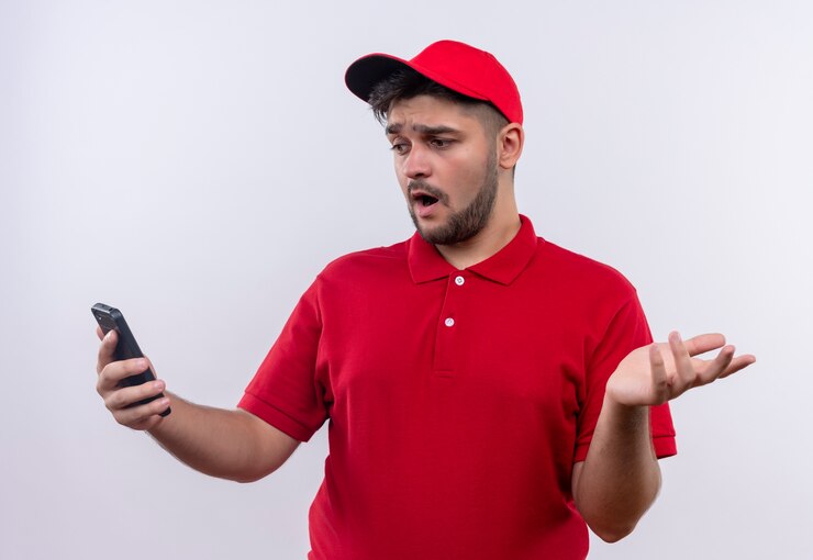 Foto gratuito repartidor joven con uniforme rojo y gorra mirando la pantalla de su teléfono inteligente con aspecto confuso e incierto