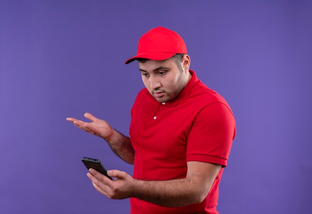 Repartidor joven con uniforme rojo y gorra mirando la pantalla de su teléfono inteligente confundido y decepcionado de pie sobre púrpura