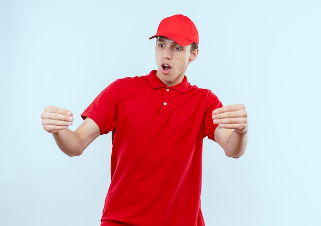 Repartidor joven en uniforme rojo y gorra mirando sorprendido gesticulando con las manos, concepto de lenguaje corporal de pie sobre una pared blanca