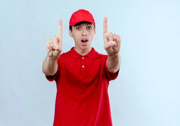 Repartidor joven en uniforme rojo y gorra mirando sorprendido mostrando los dedos índices con ambas manos de pie sobre la pared blanca