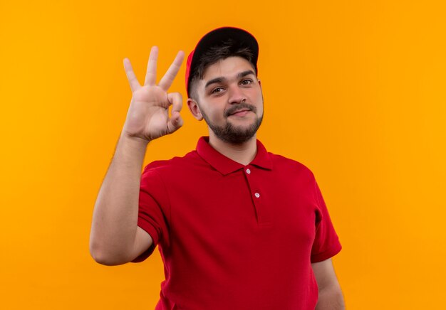 Repartidor joven en uniforme rojo y gorra mostrando signo ok sonriendo confiado