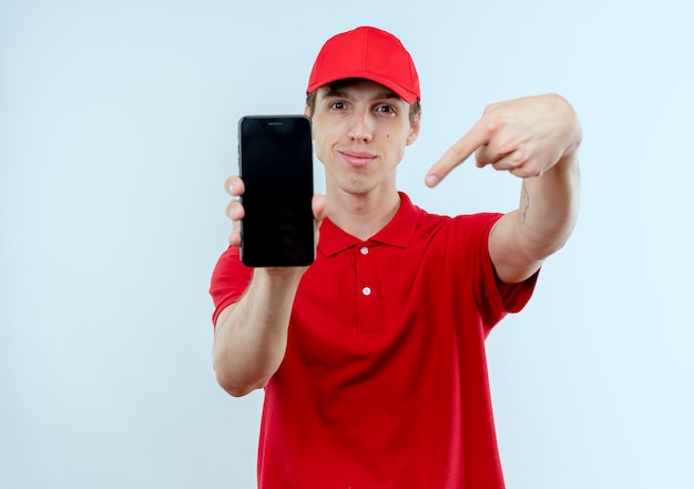 Repartidor joven en uniforme rojo y gorra mostrando smartphone apuntando con el dedo hacia él mirando confiado de pie sobre la pared blanca