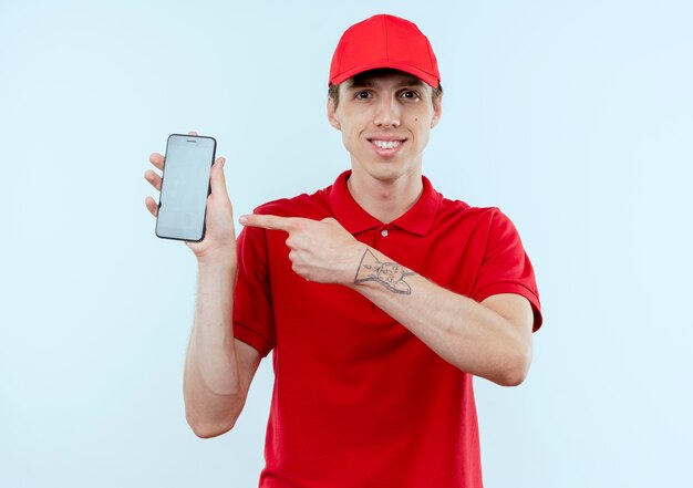 Repartidor joven en uniforme rojo y gorra mostrando smartphone apuntando con el dedo hacia él mirando confiado de pie sobre la pared blanca