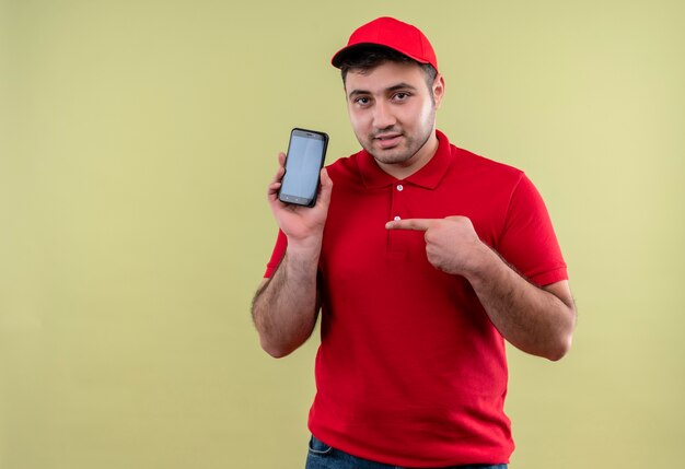 Repartidor joven en uniforme rojo y gorra mostrando smartphone apuntando con el dedo mirando confiado de pie sobre la pared verde