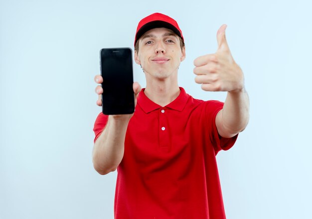 Repartidor joven en uniforme rojo y gorra mostrando smartphone sonriendo confiado mostrando los pulgares para arriba de pie sobre la pared blanca