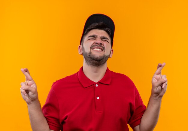 Repartidor joven en uniforme rojo y gorra con los ojos cerrados haciendo deseable cruzando los dedos con expresión de esperanza