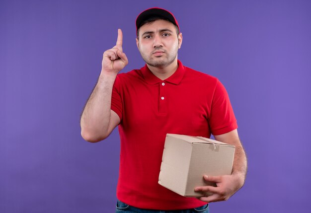 Repartidor joven en uniforme rojo y gorra con paquete de caja apuntando hacia arriba con el dedo índice con cara seria de pie sobre la pared púrpura