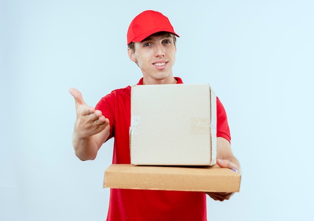 Repartidor joven en uniforme rojo y gorra con paquete de caja y caja de pizza ofreciendo con la mano al frente sonriendo de pie sobre la pared blanca