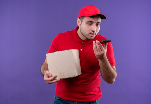 Repartidor joven en uniforme rojo y gorra con paquete de caja enviando mensaje de voz usando su teléfono inteligente parado sobre la pared púrpura
