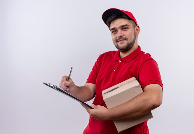 Repartidor joven en uniforme rojo y gorra con paquete de caja escribiendo en página en blanco sonriendo amigable