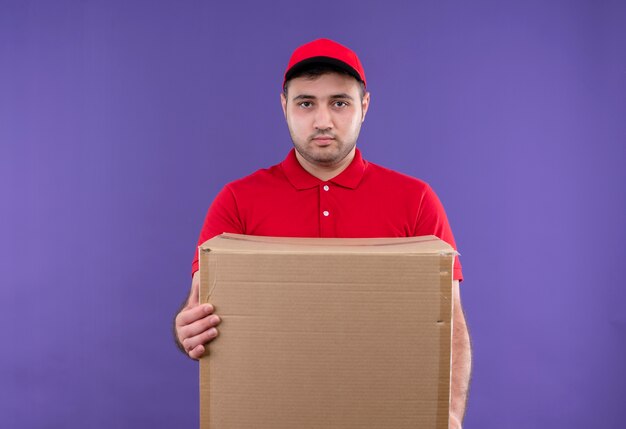 Repartidor joven en uniforme rojo y gorra con paquete de caja con expresión seria de confianza de pie sobre la pared púrpura