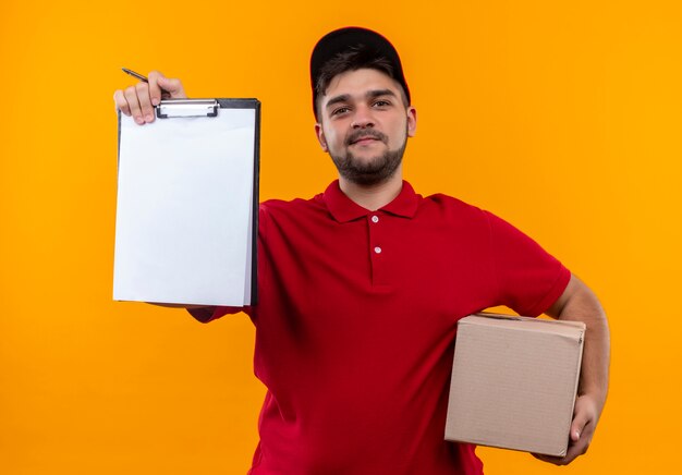 Repartidor joven en uniforme rojo y gorra con paquete de caja mostrando portapapeles con páginas en blanco pidiendo firma