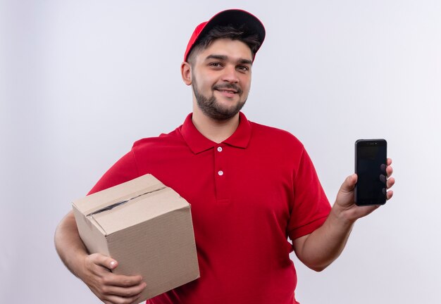 Repartidor joven en uniforme rojo y gorra con paquete de caja mostrando smartphone mirando a cámara con sonrisa segura