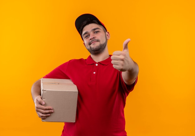 Repartidor joven en uniforme rojo y gorra con paquete de caja sonriendo confiado mostrando los pulgares para arriba
