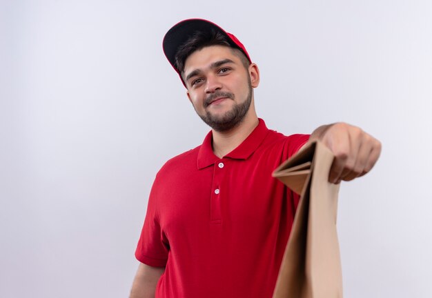 Repartidor joven en uniforme rojo y gorra con paquete de papel sonriendo amable