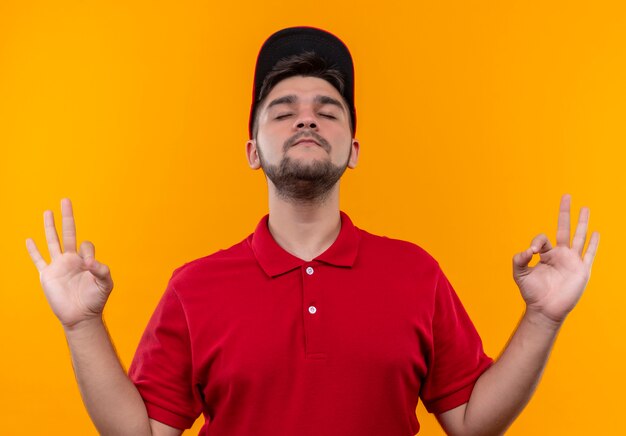 Repartidor joven en uniforme rojo y gorra relajante con los ojos cerrados haciendo gesto de meditación con los dedos