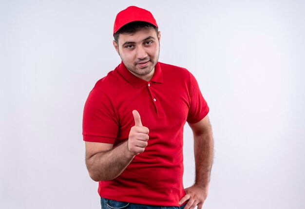 Repartidor joven en uniforme rojo y gorra sonriendo alegremente mostrando los pulgares para arriba de pie sobre la pared blanca