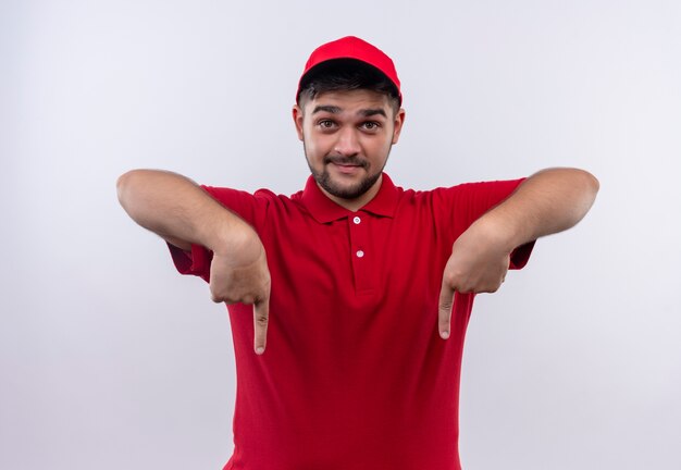 Repartidor joven en uniforme rojo y gorra sonriendo confiado apuntando con los dedos hacia abajo