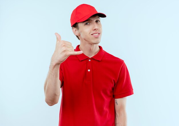 Repartidor joven en uniforme rojo y gorra sonriendo confiado haciendo gesto de llamarme de pie sobre la pared blanca