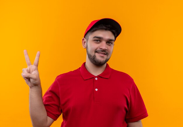 Repartidor joven en uniforme rojo y gorra sonriendo mostrando y apuntando hacia arriba con los dedos número dos