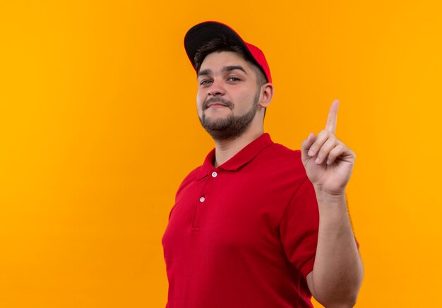 Repartidor joven en uniforme rojo y gorra sonriendo seguro mostrando el dedo índice