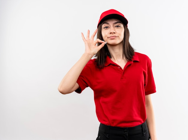 Repartidor joven con uniforme rojo y gorra con sonrisa escéptica que muestra gesto de silencio como cerrar la boca con una cremallera de pie sobre una pared blanca