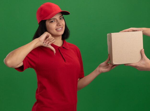 Repartidor joven en uniforme rojo y gorra sosteniendo una caja de cartón a un cliente que parece disgustado mostrando los pulgares hacia abajo de pie sobre la pared verde