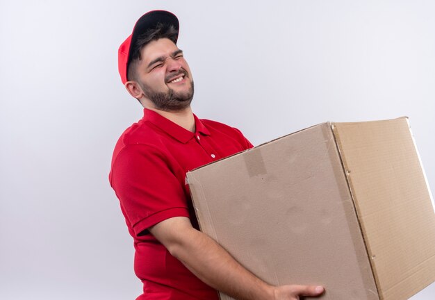 Repartidor joven en uniforme rojo y gorra sosteniendo una caja de cartón grande que sufre de peso pesado