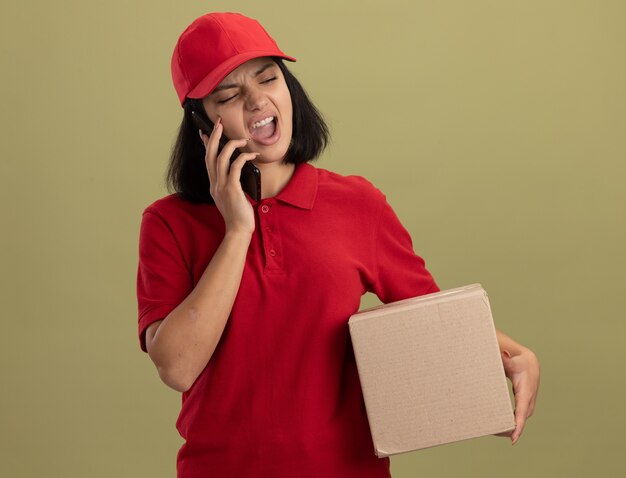 Repartidor joven en uniforme rojo y gorra sosteniendo una caja de cartón gritando mientras habla por teléfono móvil de pie sobre una pared de luz