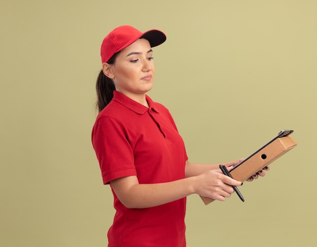 Repartidor joven en uniforme rojo y gorra sosteniendo caja de pizza con portapapeles y lápiz mirando confiado parado sobre pared verde