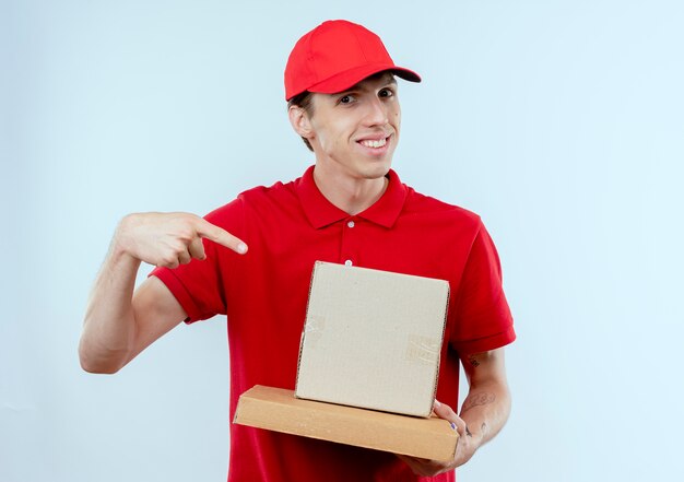 Repartidor joven en uniforme rojo y gorra sosteniendo cajas de cartón apuntando con el dedo índice a ellos sonriendo confiado de pie sobre la pared blanca