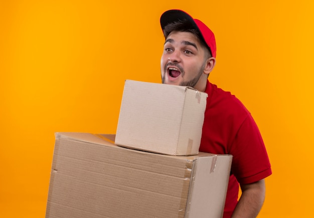 Repartidor joven en uniforme rojo y gorra sosteniendo cajas de cartón mirando sorprendido