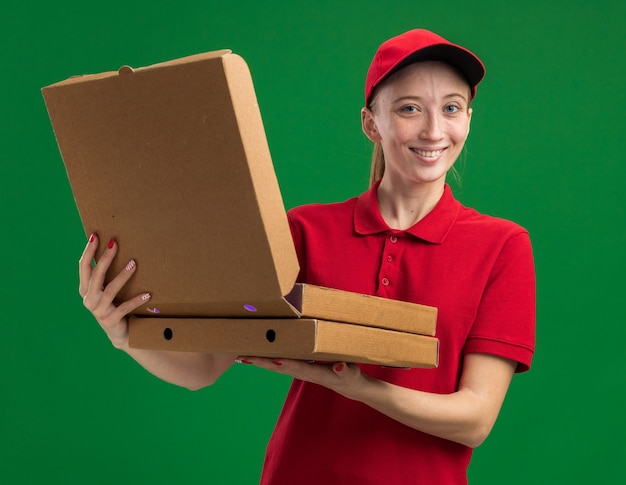 Repartidor joven en uniforme rojo y gorra sosteniendo cajas de pizza abriendo uno de ellos sonriendo amistoso de pie sobre la pared verde