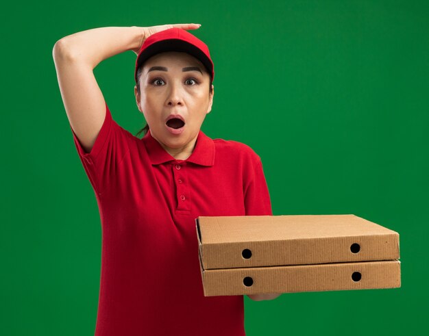 Repartidor joven en uniforme rojo y gorra sosteniendo cajas de pizza asombrado con la mano en la cabeza de pie sobre la pared verde