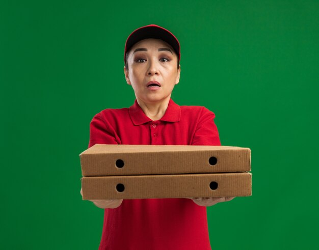 Repartidor joven en uniforme rojo y gorra sosteniendo cajas de pizza confundidas