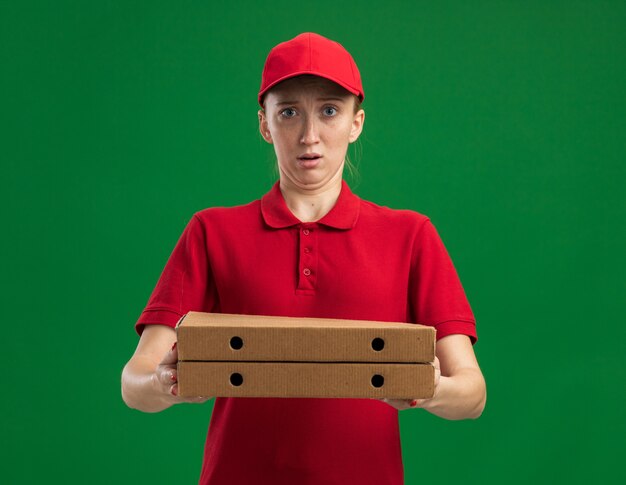 Repartidor joven en uniforme rojo y gorra sosteniendo cajas de pizza confundido y sorprendido de pie sobre la pared verde