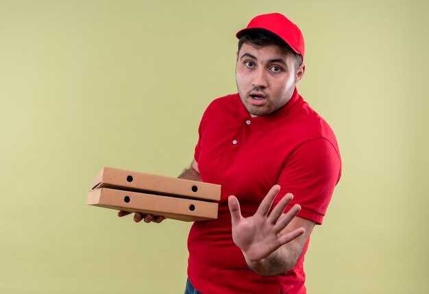 Repartidor joven en uniforme rojo y gorra sosteniendo cajas de pizza haciendo señal de pare con la mano con expresión de miedo de pie sobre la pared verde