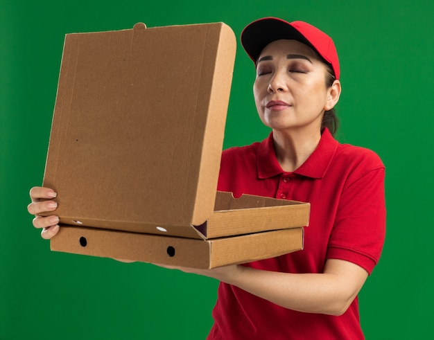 Repartidor joven en uniforme rojo y gorra sosteniendo cajas de pizza inhalando un aroma agradable con los ojos cerrados de pie sobre la pared verde