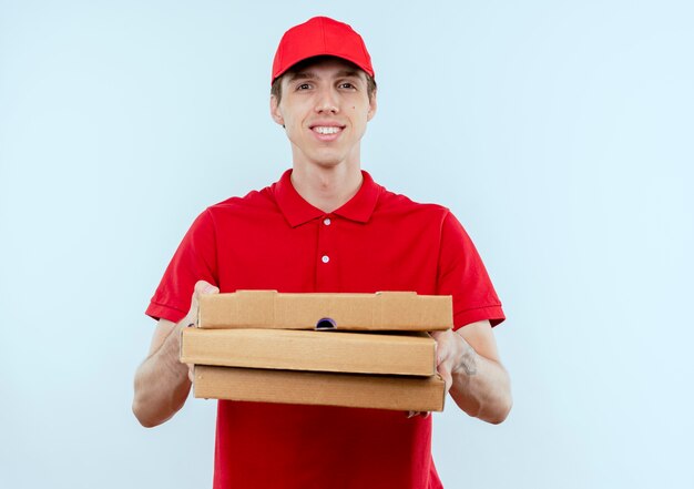 Repartidor joven en uniforme rojo y gorra sosteniendo cajas de pizza mirando al frente con expresión de confianza de pie sobre la pared blanca