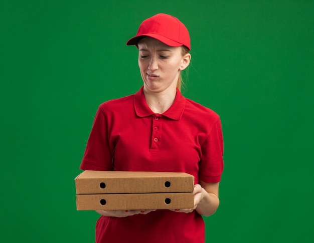 Repartidor joven en uniforme rojo y gorra sosteniendo cajas de pizza mirándolos confundidos y disgustados de pie sobre la pared verde