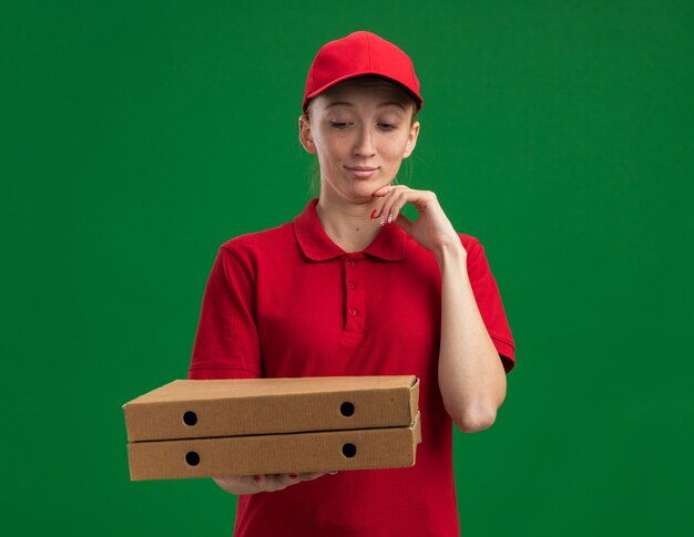 Repartidor joven en uniforme rojo y gorra sosteniendo cajas de pizza mirándolos con la mano en la barbilla pensando de pie sobre la pared verde
