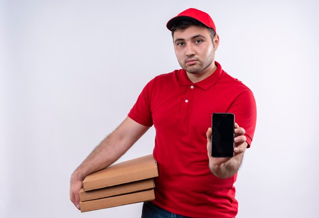 Repartidor joven en uniforme rojo y gorra sosteniendo cajas de pizza mostrando smartphone con expresión de confianza de pie sobre la pared blanca