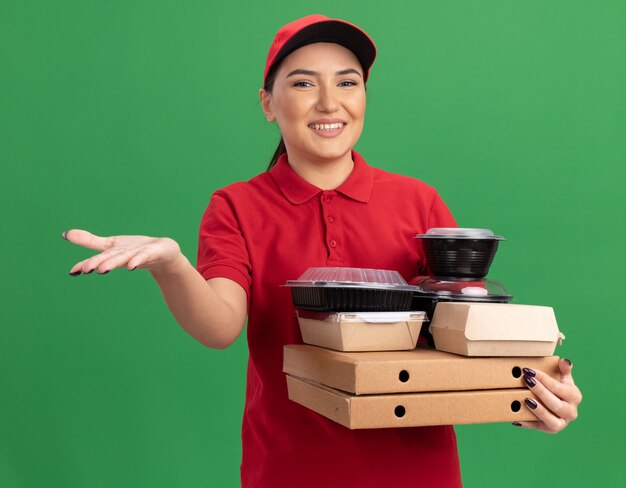 Repartidor joven en uniforme rojo y gorra sosteniendo cajas de pizza y paquetes de alimentos mirando al frente con una sonrisa en la cara con el brazo levantado haciendo un gesto de bienvenida de pie sobre una pared verde