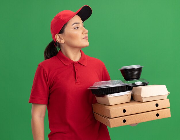 Repartidor joven en uniforme rojo y gorra sosteniendo cajas de pizza y paquetes de alimentos mirando a un lado con cara seria de pie sobre la pared verde