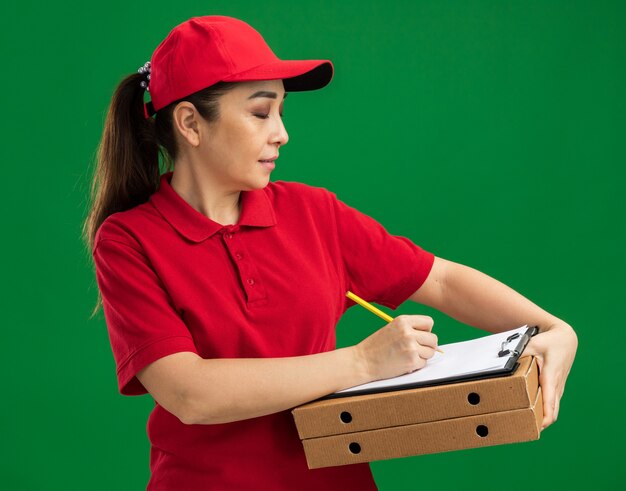 Repartidor joven en uniforme rojo y gorra sosteniendo cajas de pizza y portapapeles con bolígrafo escribiendo algo con expresión de confianza