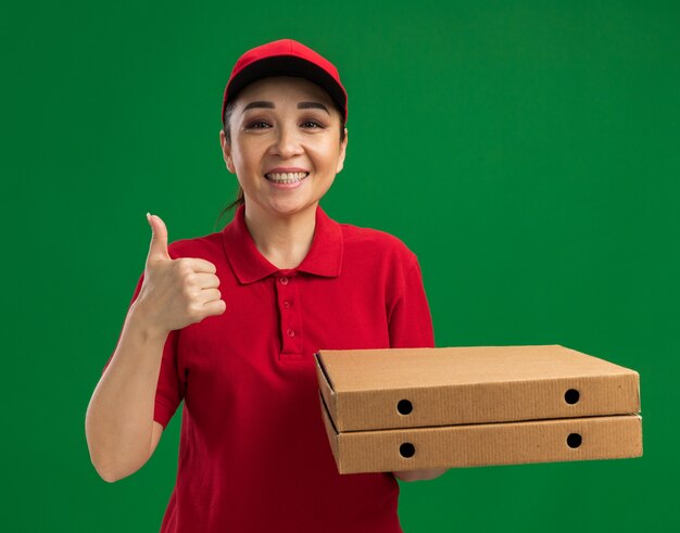 Repartidor joven en uniforme rojo y gorra sosteniendo cajas de pizza sonriendo amigable mostrando los pulgares para arriba de pie sobre la pared verde