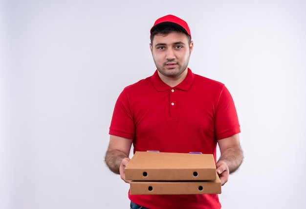 Repartidor joven en uniforme rojo y gorra sosteniendo cajas de pizza sonriendo confiado de pie sobre la pared blanca