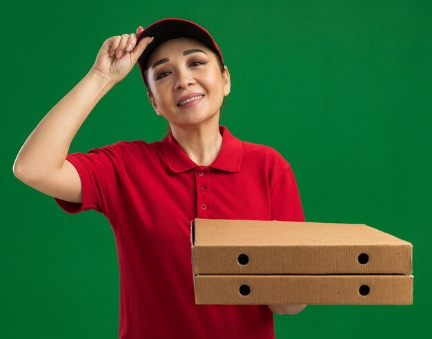 Repartidor joven en uniforme rojo y gorra sosteniendo cajas de pizza sonriendo confiado