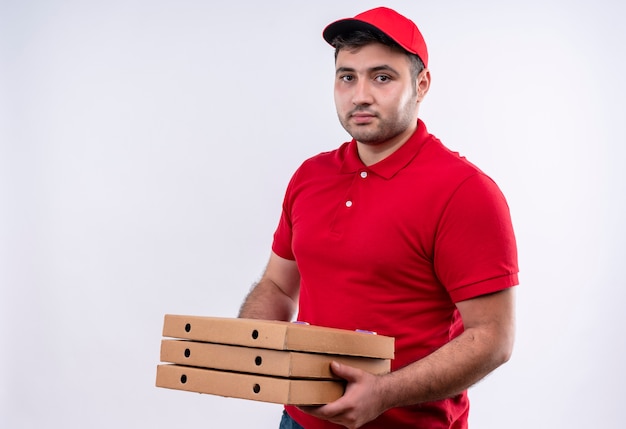 Repartidor joven en uniforme rojo y gorra sosteniendo cajas de pizza sonriendo con expresión de confianza de pie sobre la pared blanca